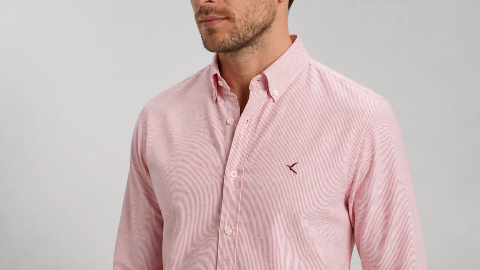 Close studio view of a pale pink shirt showing its fine stripe texture, button-down collar and small pocket-region logo